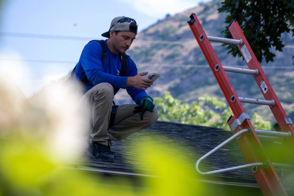 BRS-Installer-on-Roof-in-Bright-Blue-Shirt Male with hat backwards, squatting on a roof searching solar panel options on his smartphone