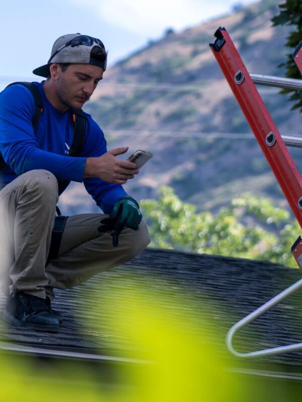 Male with hat backwards, squatting on a roof looking at a smartphone with ladder next to him