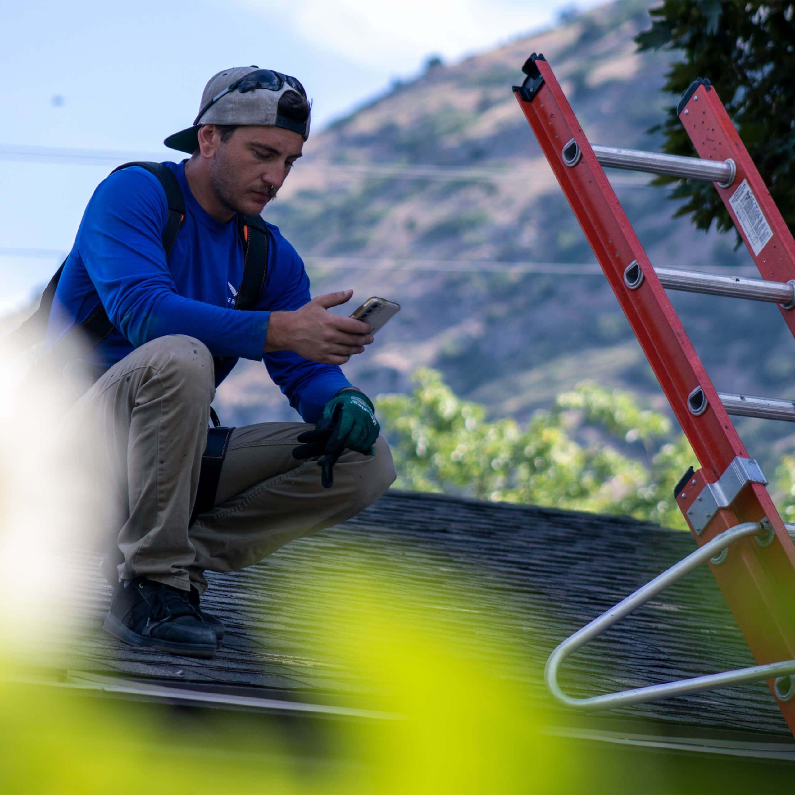Male with hat backwards, squatting on a roof looking at a smartphone with ladder next to him