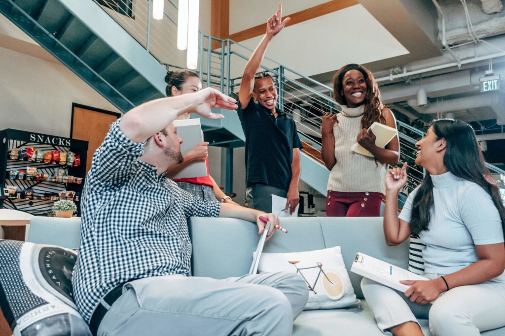 Casual-Team-Meeting Two individuals sitting on a couch, with a small group of people in the background - casually working together