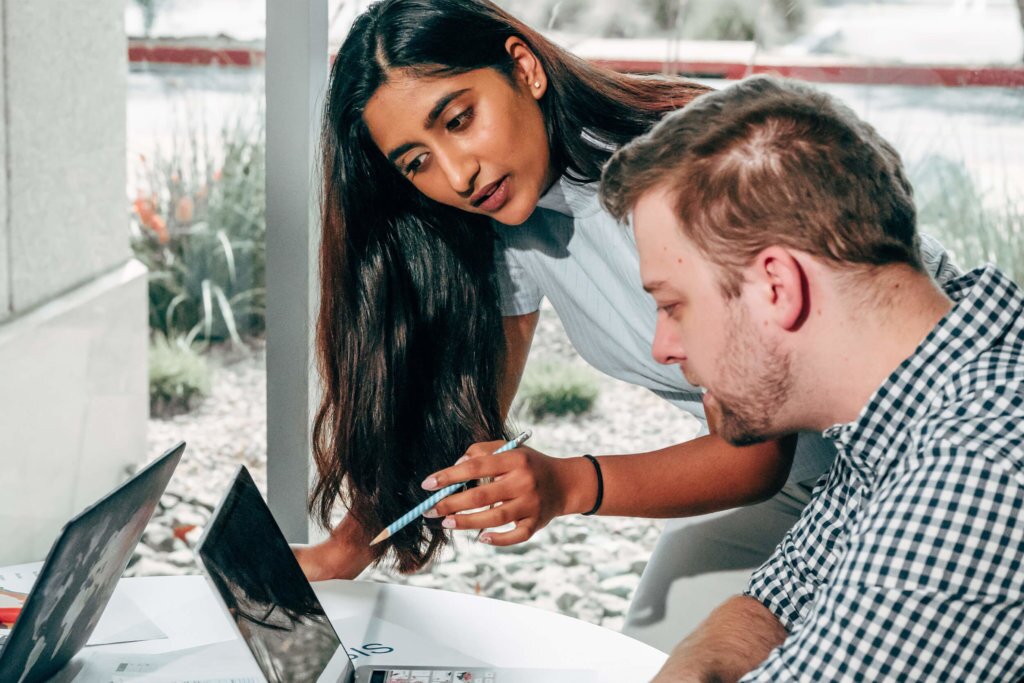 Two-Employees-Working-Together Young, female instructing co-worker in a plaid shirt working on a laptop