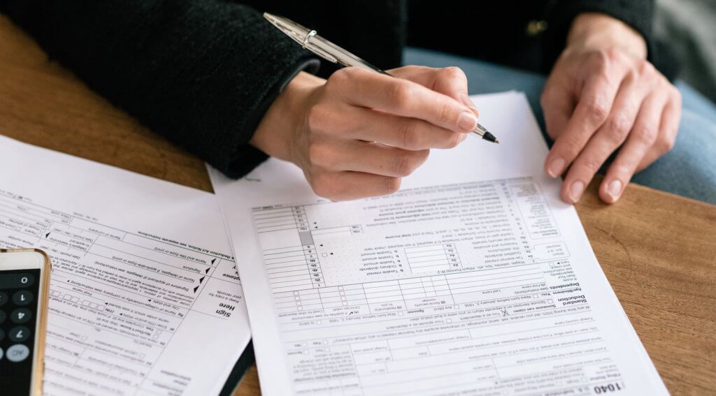 An individual filing tax paper and paperwork on a deak, using their phone as a calculator