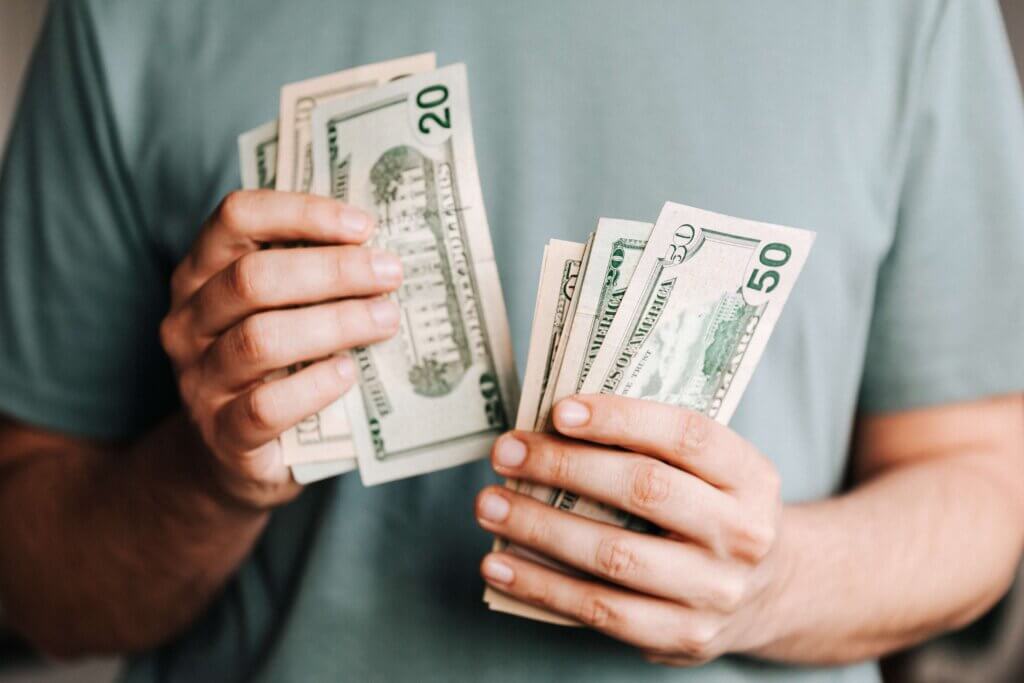 Male in sage green shirt holding large US dollar bills in both hands, fanned out