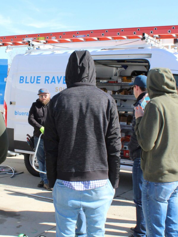 Small group of Blue Raven Solar employees on-site listening to instructions, in a semi-circle with a company branded install van in the background