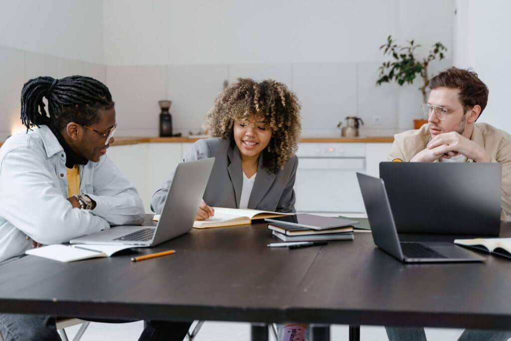 Three employees, gathered around a conference table with laptops and notebooks talking to one another