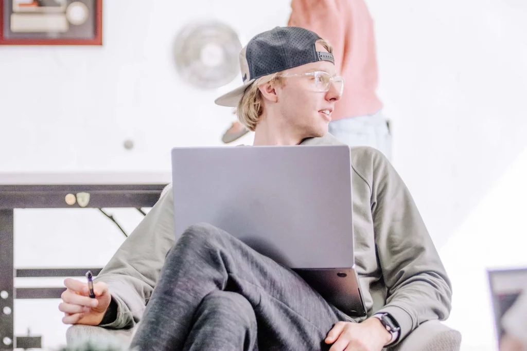 Blue-Raven-Solar-Employee-Working-on-Laptop Young male, hat backwards, and glasses working on a laptop looking to the right