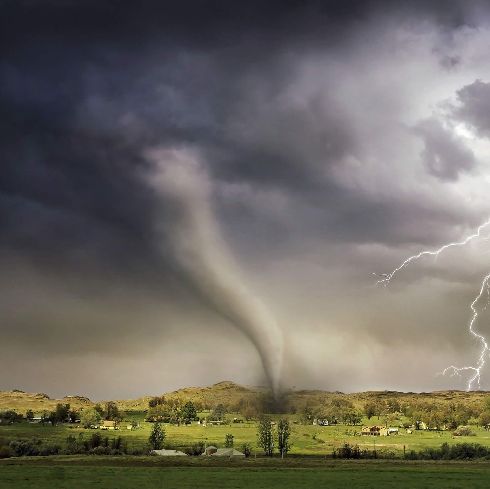 Dark clouds, tornado, and lightening striking a green hill scattered with houses and trees