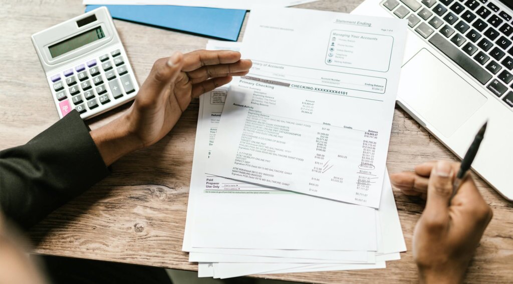 Black male preparing personal tax documents by looking through bank statements, using calculator, and looking up information on laptop