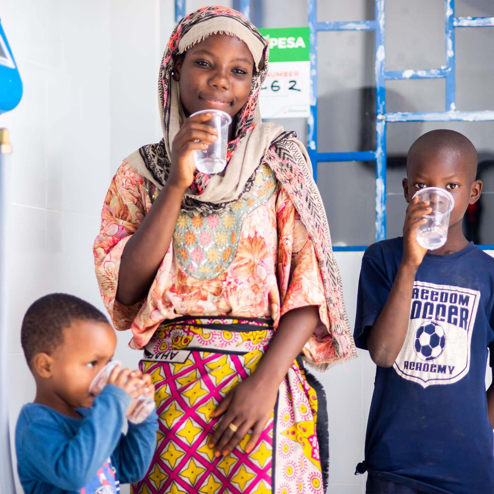 Young female drinking water out of a clear cup, surrounded by two little boys also drinking water out of cups