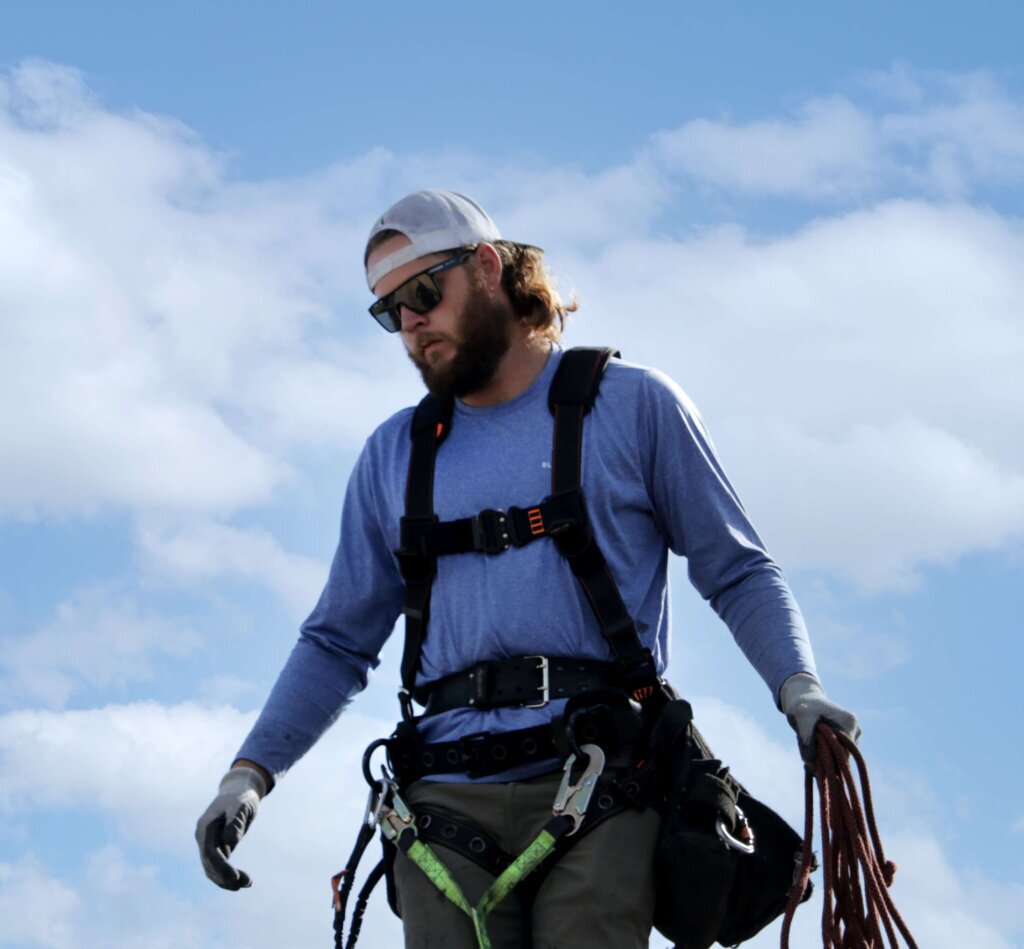 Blue Raven Solar Installer-cropped Blue Raven Solar installer in a harness, carrying specialized rope and equipment with blue skies and clouds behind