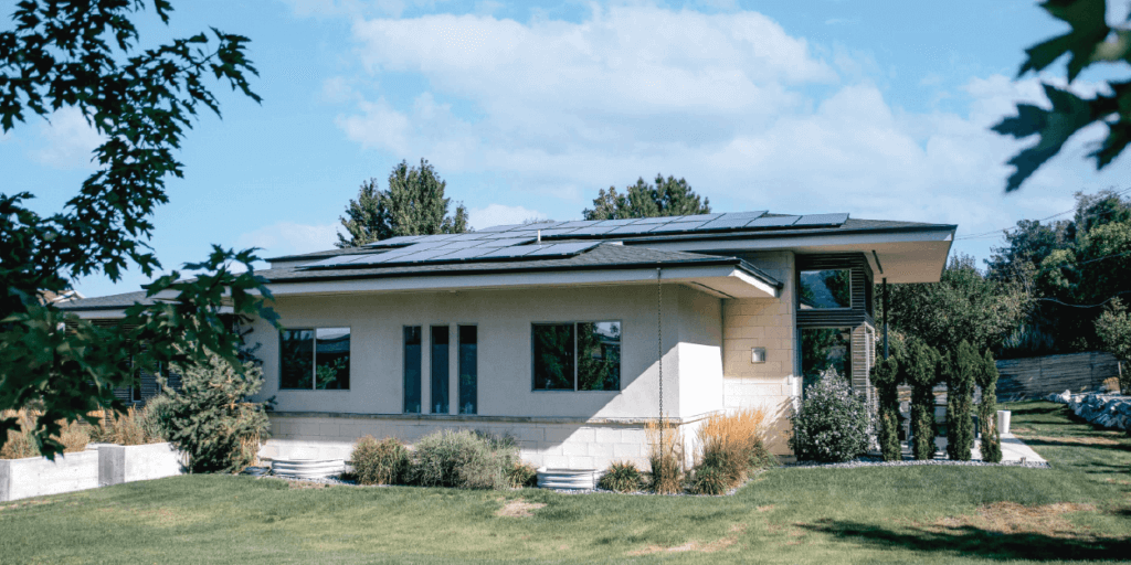Neutral exterior home with large solar panel system on roof, surrounded by trees and baby blue sky above