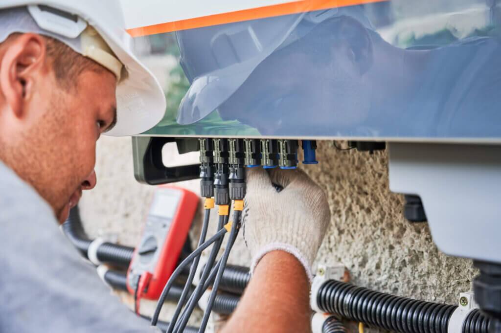 Male in a hardhat and gloves working on electrical systems mounted on the side of a structure and connecting cords to ensure proper operation