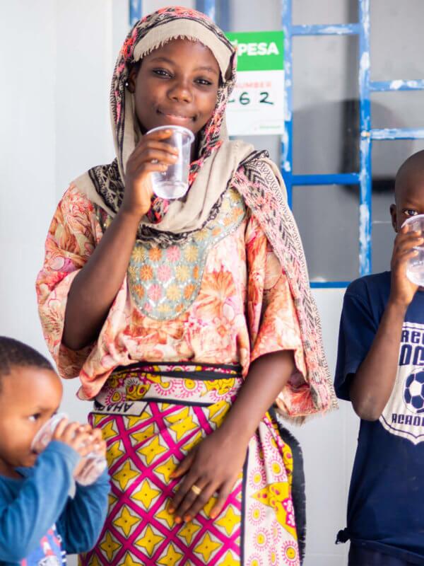 Young female drinking water out of a clear cup, surrounded by two little boys also drinking water out of cups