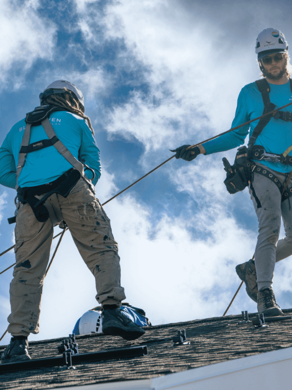 Two Blue Raven Solar installers in teal branded t-shirts utilizing specialized equipment to secure solar panels to a homeowner's roof