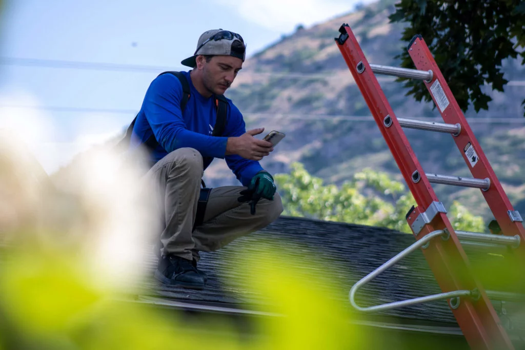 Male with hat backwards, squatting on a roof searching solar panel options on his smartphone