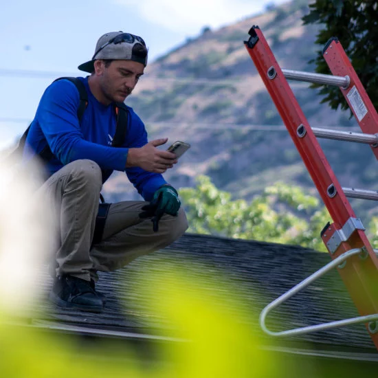 Male with hat backwards, squatting on a roof looking at a smartphone with ladder next to him