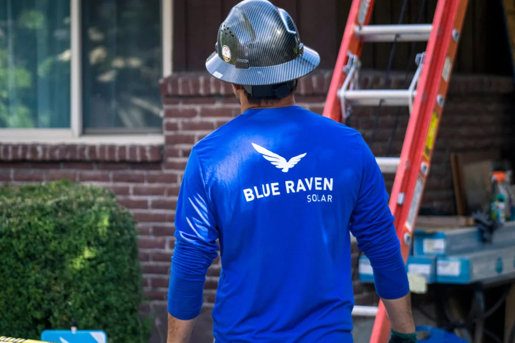 Blue Raven Solar Installer in Blue Shirt Blue Raven Solar installer in blue branded long-sleeve shirt and hardhat on, walking towards a ladder leading to the roof of a house
