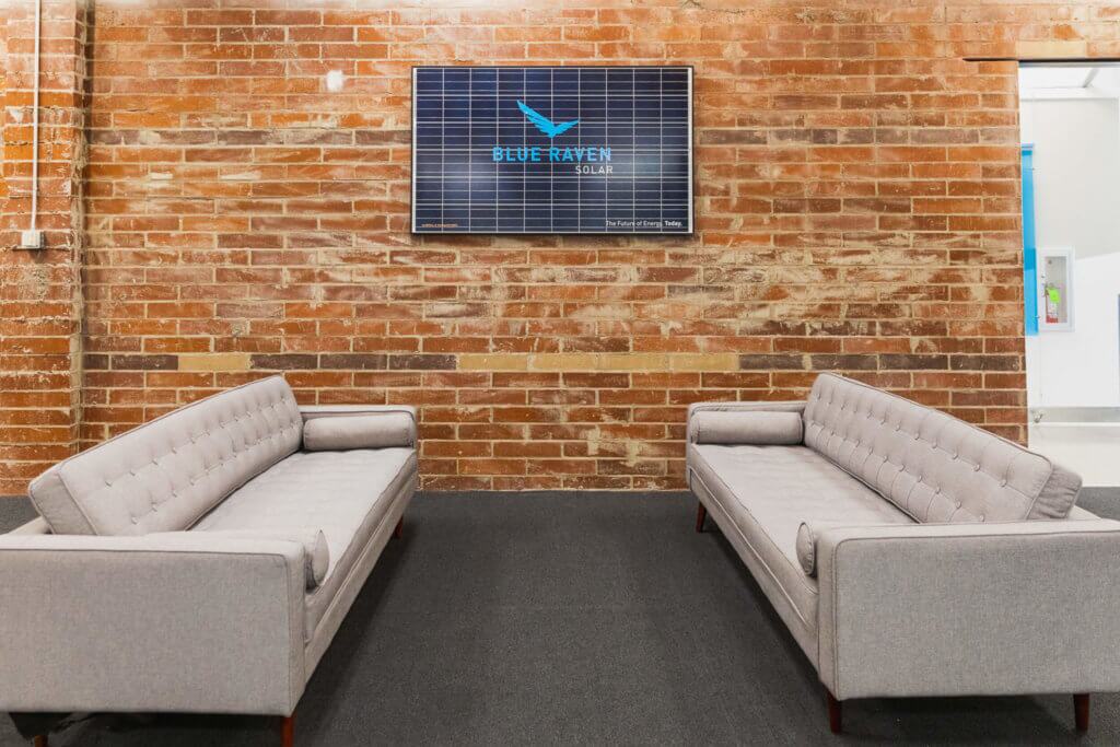 Blue-Raven-Solar-Corporate-Office-Staged Blue Raven Solar corporate office featuring two grey couches in front of an exposed brick wall and the logo mounted on a solar panel