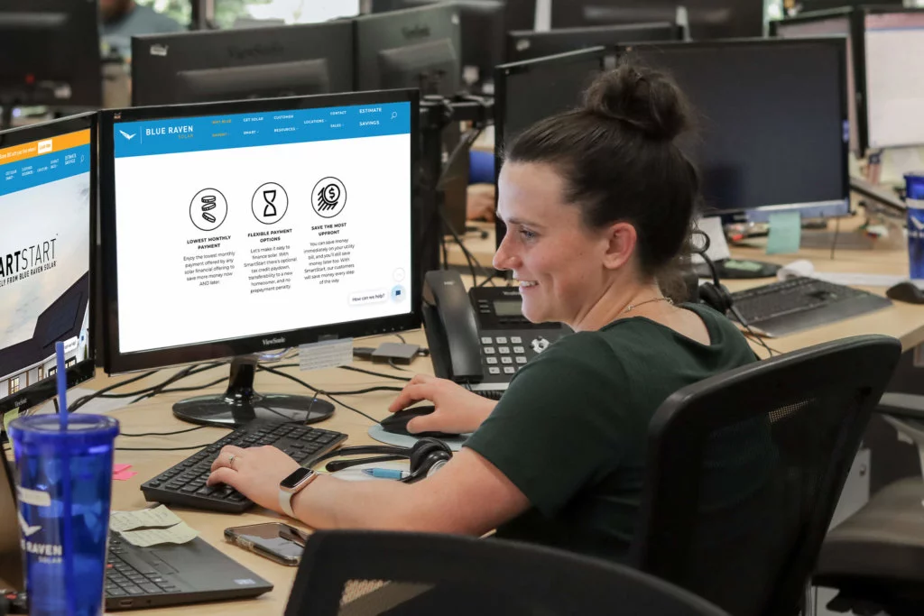 Blue-Raven-Solar-Employee-Sitting-at-Computer Female in a dark green short sleeve shirt and with her hair in a bun, sitting and working on two computer monitors