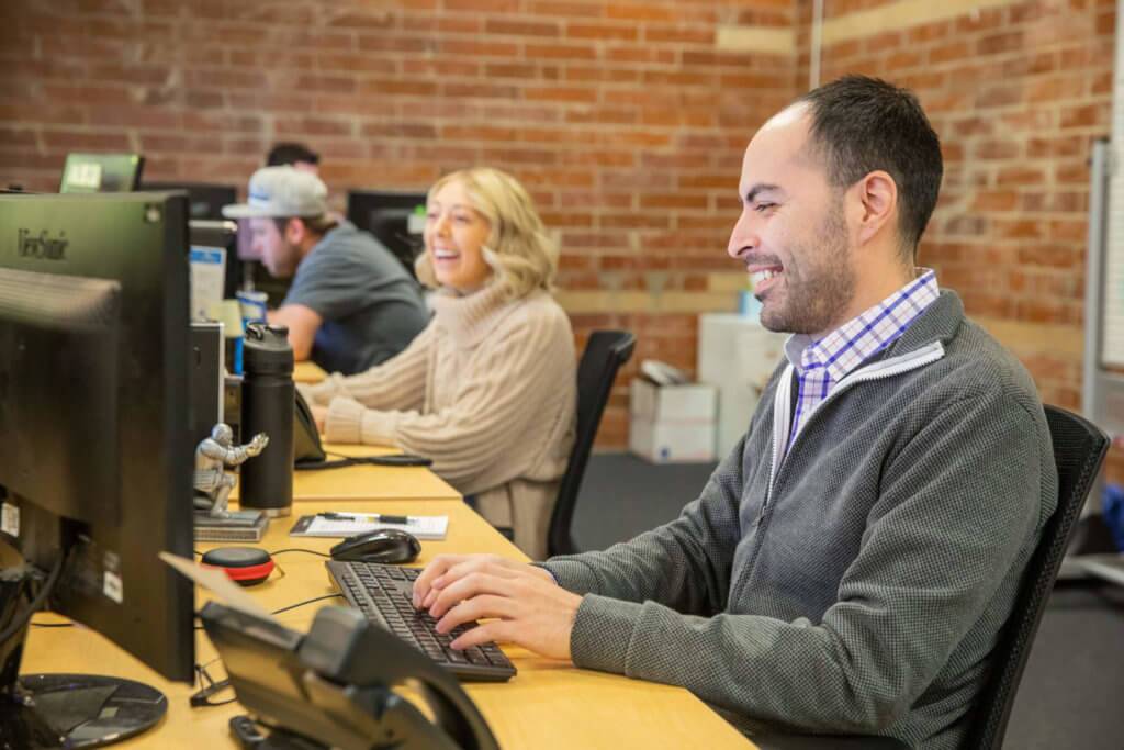 Three-Blue-Raven-Solar-Employees-Sitting-at-Their-Desks Three Blue Raven Solar employees sitting at their desks, smiling and working