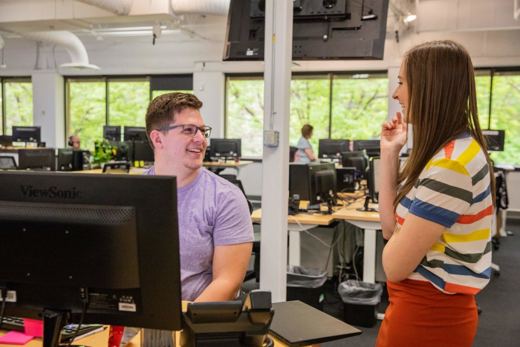 Two-Blue-Raven-Solar-Employees-Discussing A male and female dicussing with each other in an open-floor office building, surrounded by computer monitors and desks