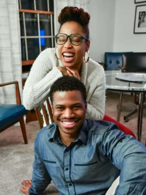 A black couple, male sitting on the floor smiling and the female sitting in a chair laughing with her eyes closed