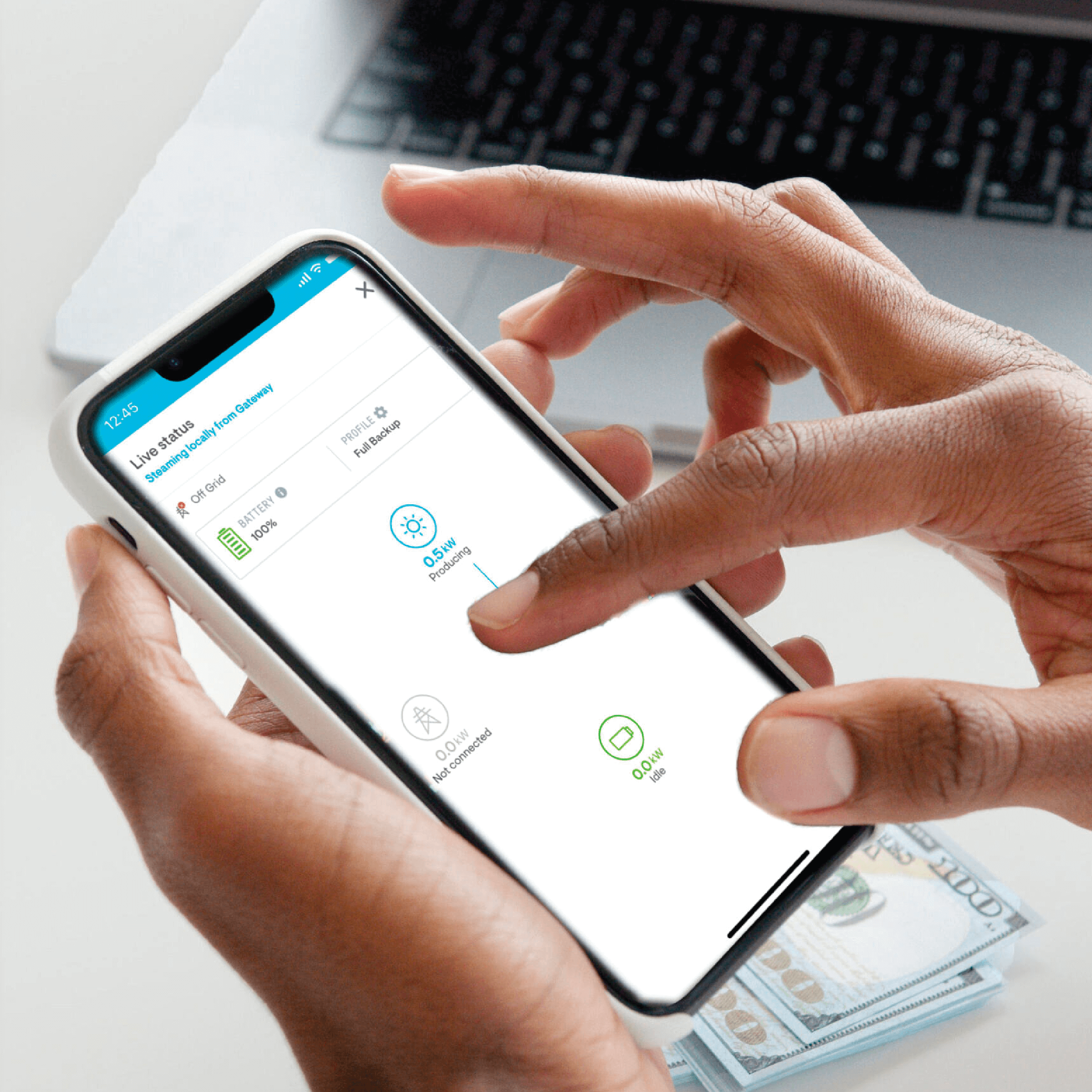 Mobile device held between two hands, scrolling with cash and a laptop in the background