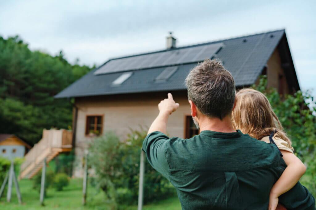 Father in a green shirt, holding a small child and pointing to a large structure with a row of solar panels on the roof