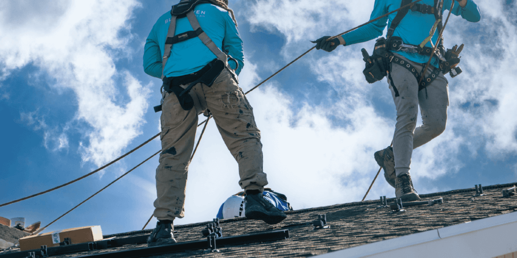 Two Blue Raven Solar installers in teal branded t-shirts utilizing specialized equipment to secure solar panels to a homeowner's roof