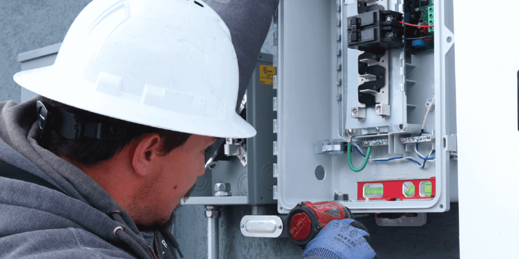 Electrician with a white hard hat, securing a meter to the side of a house using power tools