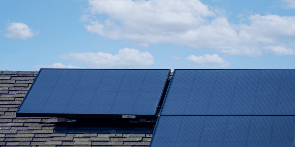 Benefits of Investing in Solar Battery Storage-02 Close-up shot of a 3-panel solar array on grey asphalt shingles, with a light blue sky and clouds in the background